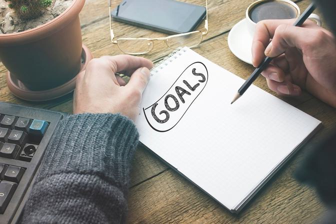 Person writing GOALS on notebook at wooden desk with laptop, keyboard, coffee mug, and phone, overhead