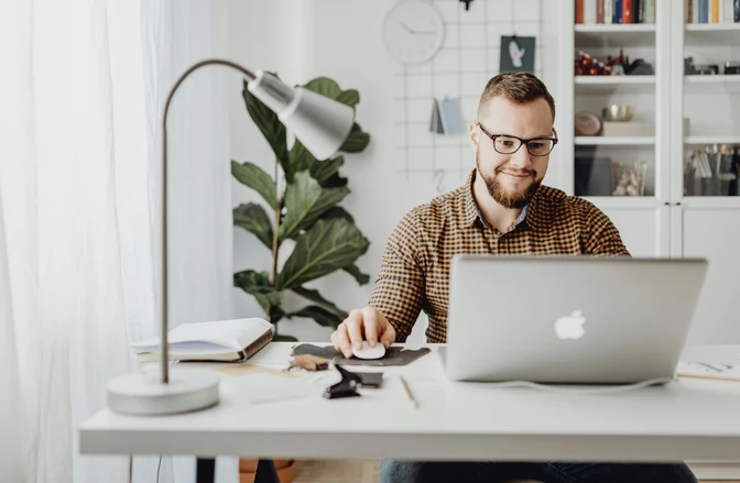 Man in plaid shirt and glasses working at white desk with laptop, arc lamp, potted plant, and shelving in background