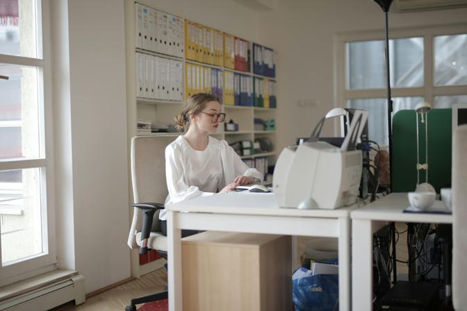 Woman with glasses typing at white desk in bright modern office with shelving, natural light from windows