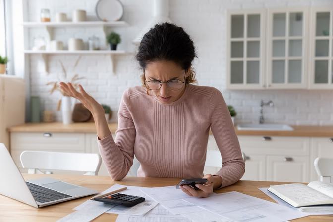 Woman in glasses and pink ribbed sweater at wooden kitchen desk with laptop, calculator, and papers, frustrated expression