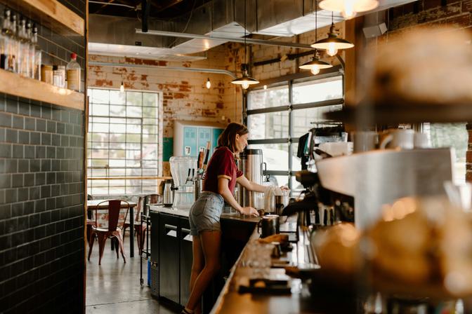 Barista in maroon shirt working behind espresso machine in industrial cafe interior, brick walls, pendant lights