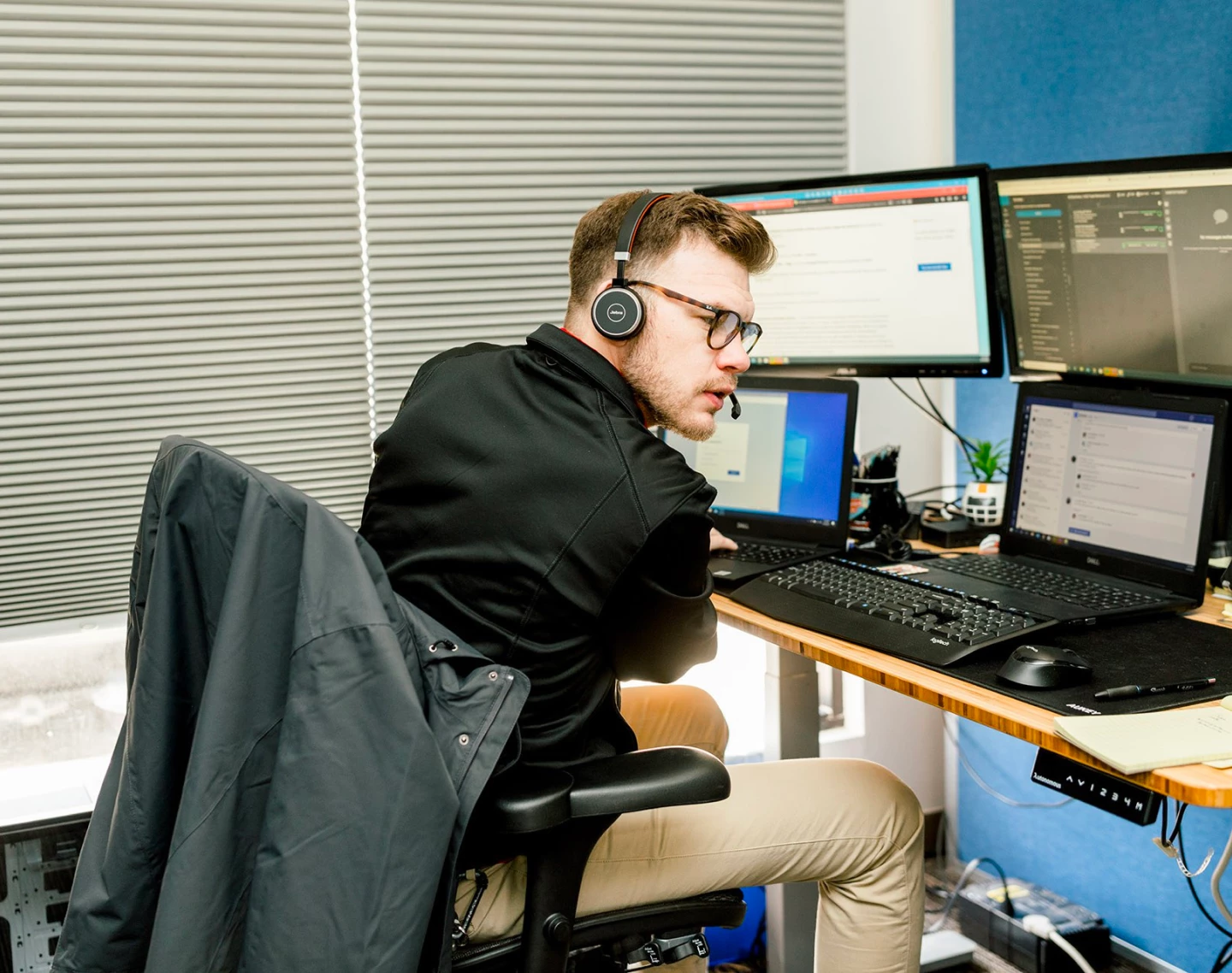 Man wearing headset and glasses at wooden desk with multiple monitors and laptop