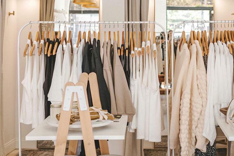 Retail clothing rack with wooden hangers holding white, black, and beige garments in a bright showroom with natural windows