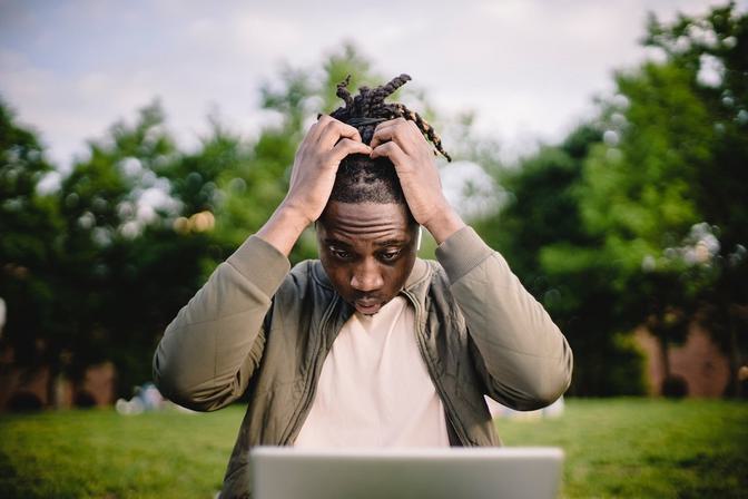 Young man sitting outdoors with a green tree background, holding a potted plant above his head while working at a laptop