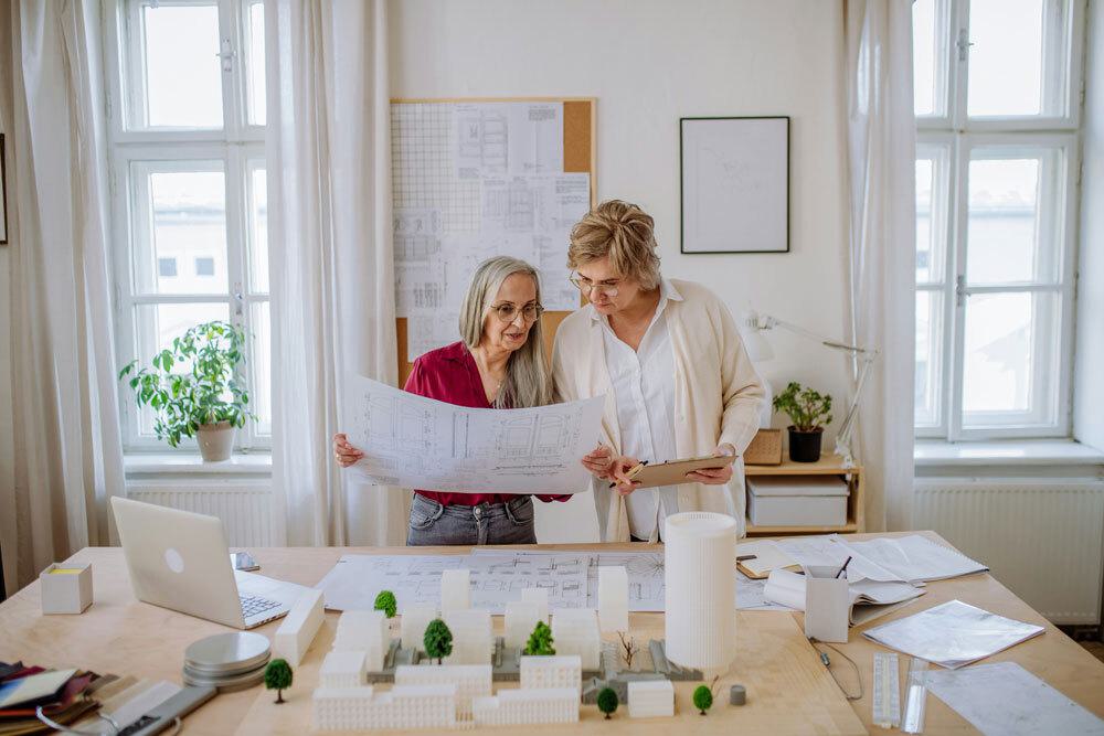Two professional women reviewing architectural blueprint in bright office workspace with architectural model, laptop
