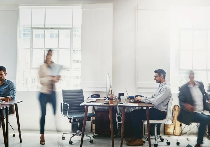 Busy open-plan office with multiple employees at desks, motion blur of moving people, natural light from large windows