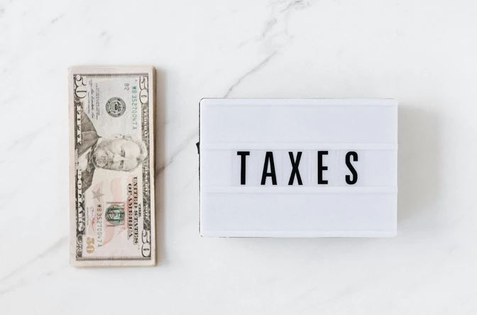 Dollar bill and lightbox with 'TAXES' text on marble surface, flat lay product shot, financial services concept