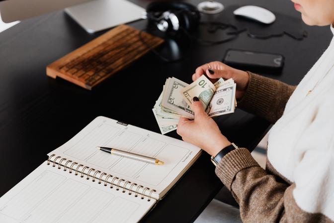 Person in brown cardigan holding British pound notes over open spiral notebook on black desk, workspace with laptop, camera